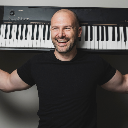 A person smiling with arms raised next to a pianoDescription automatically generated